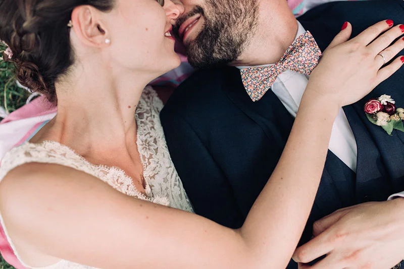 Gros plan romantique de Clara et Julien s'embrassant, elle en robe de dentelle et lui en costume bleu et nœud papillon.