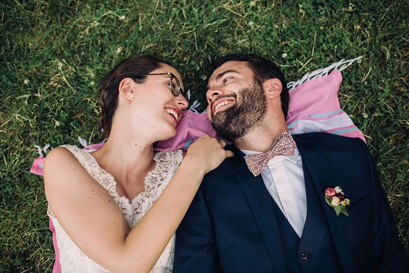 Jeunes mariés souriants allongés sur l'herbe, Clara en robe blanche et Julien en costume bleu avec un nœud papillon.