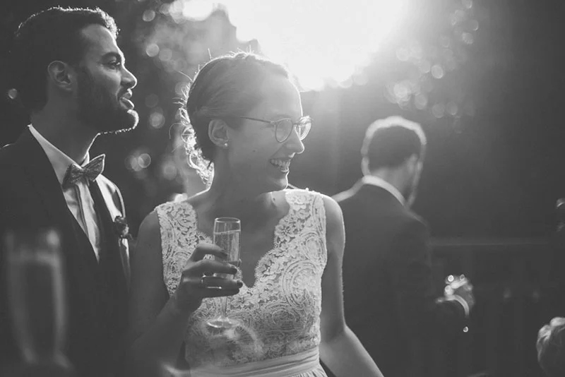 Couple souriant en tenue de mariage, photo noir et blanc lumineuse, la mariée tenant une flûte de champagne.