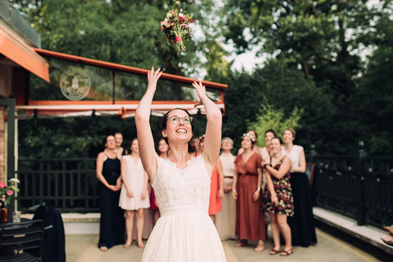 Une mariée en robe blanche lance son bouquet à un groupe d'invitées lors d'une réception en extérieur.