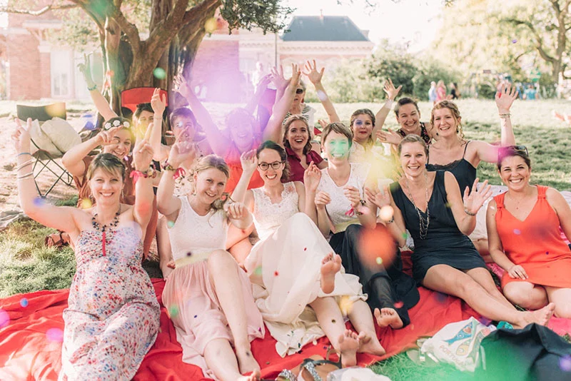 Groupe de femmes souriantes et festives assises sur une nappe rouge dans un jardin ensoleillé avec des effets de lumière.