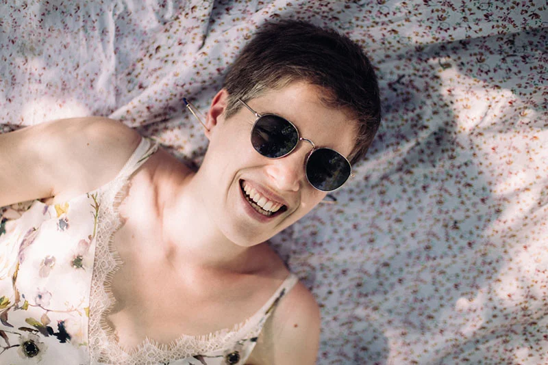 Une femme souriante aux cheveux courts et lunettes de soleil, allongée sur un drap floral dans une ambiance lumineuse.