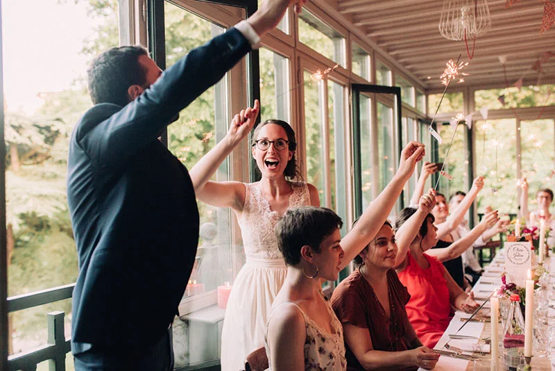 Jeunes mariés et invités célébrant avec des cierges magiques lors d'un dîner festif et lumineux en intérieur.