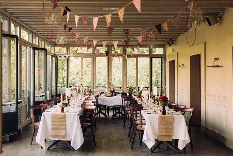 Salle de réception lumineuse avec tables dressées, guirlandes de fanions colorés et ambiance champêtre et romantique.
