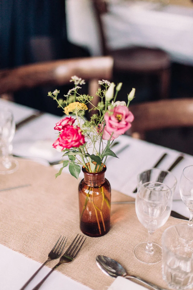 Petit bouquet de fleurs champêtres roses et jaunes dans un vase ambré sur une table de mariage élégante et naturelle.