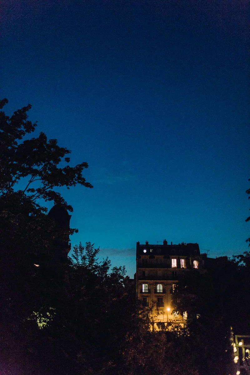 Vue nocturne d'un immeuble parisien éclairé sous un ciel bleu profond, entouré de silhouettes d'arbres sombres.