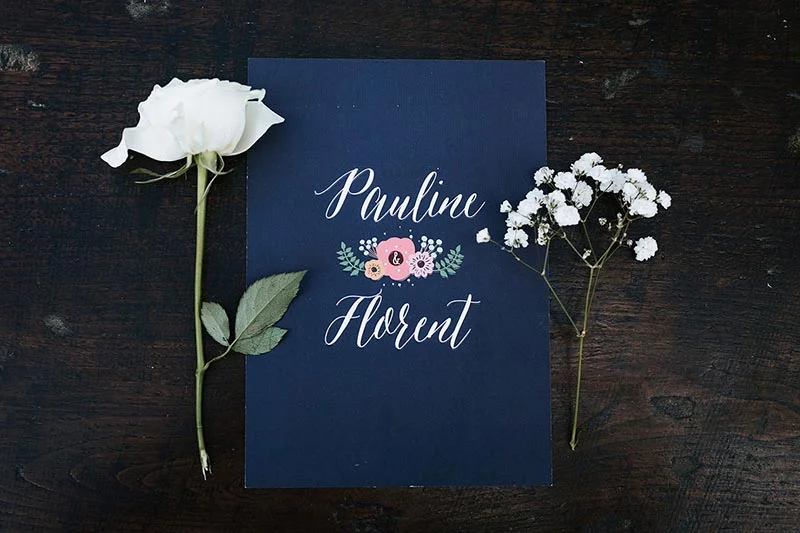 Faire-part bleu marine avec les prénoms Pauline et Florent, entouré d'une rose blanche et de gypsophile sur fond bois.