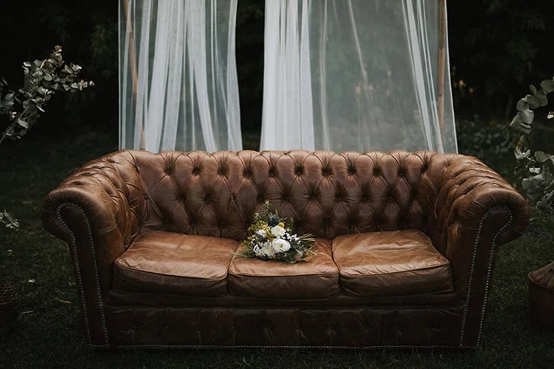Canapé Chesterfield en cuir marron avec un bouquet de fleurs blanches posé dessus, devant un rideau blanc vaporeux.