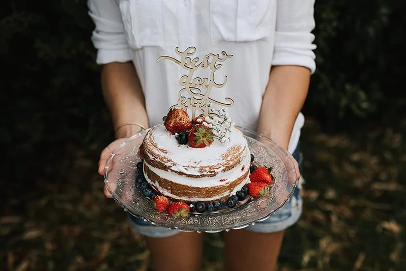 Une personne tenant un gâteau nu aux fraises et myrtilles avec un cake topper doré sur fond de nature sombre.