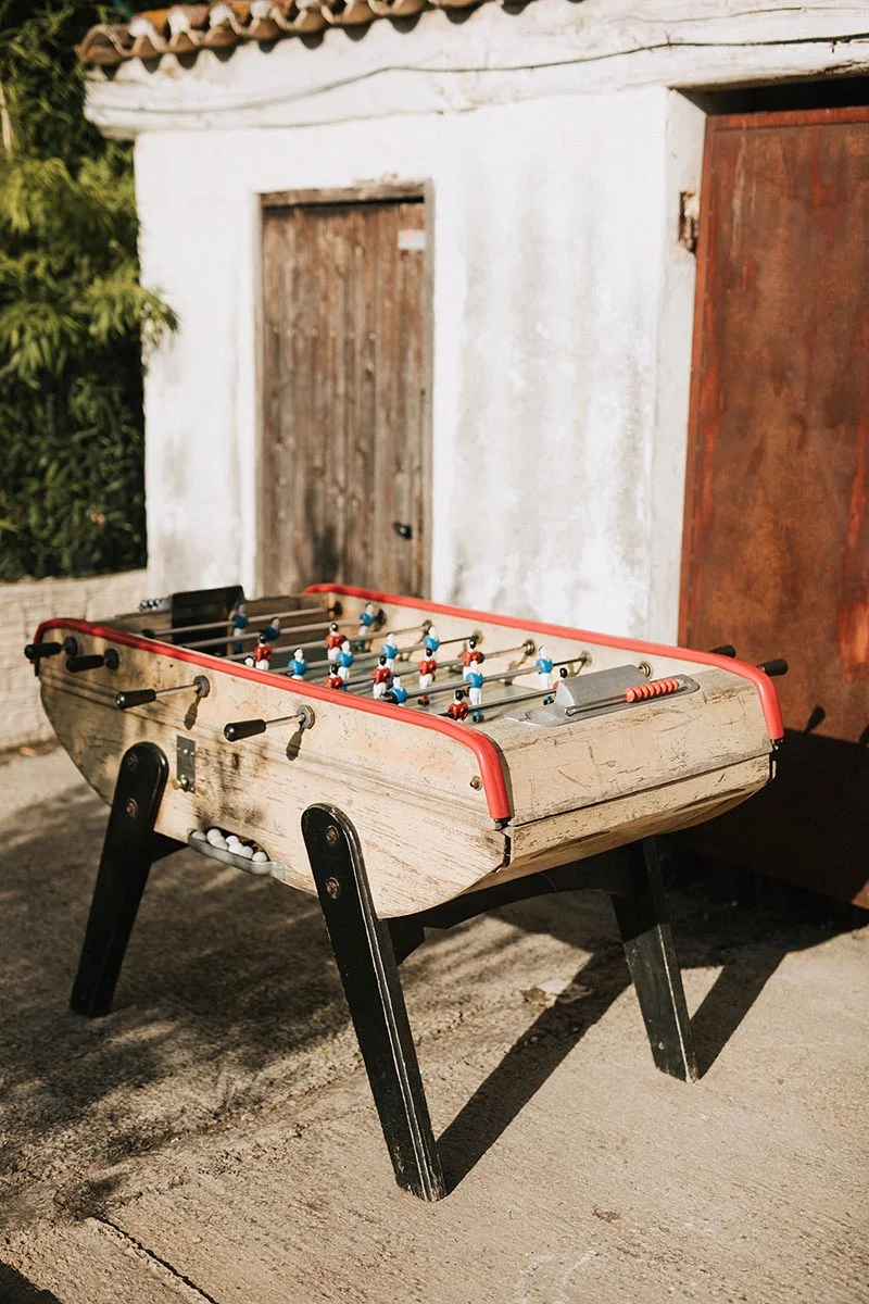 Baby-foot vintage en bois installé en extérieur devant un mur blanc et une porte rustique.