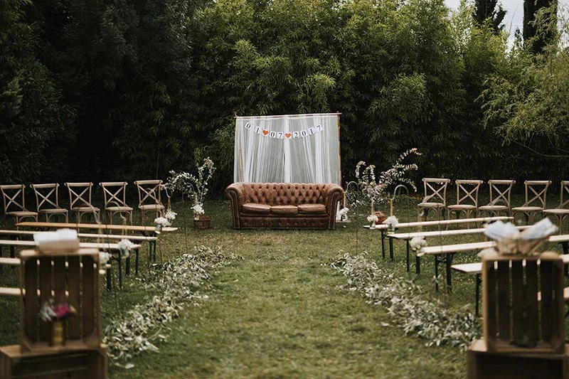 Décoration de cérémonie en plein air avec canapé Chesterfield brun, rideau blanc et allée de feuilles sur gazon.