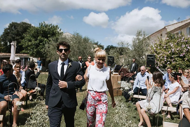 Un couple souriant marche bras dessus bras dessous lors d'une cérémonie de mariage en plein air et ensoleillée.