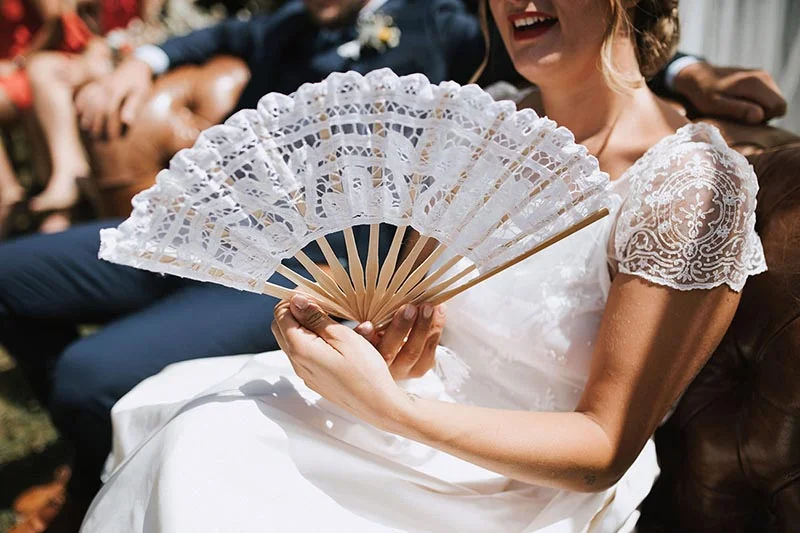 Une mariée souriante en robe blanche tenant un éventail en dentelle blanche lors de son mariage.