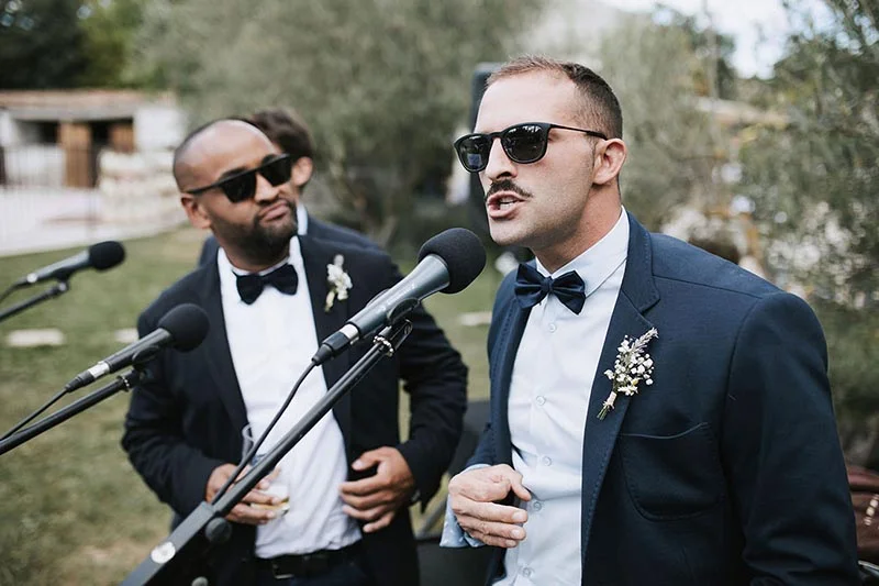 Deux hommes en costume bleu marine et nœud papillon portant des lunettes de soleil lors d'un discours de mariage.