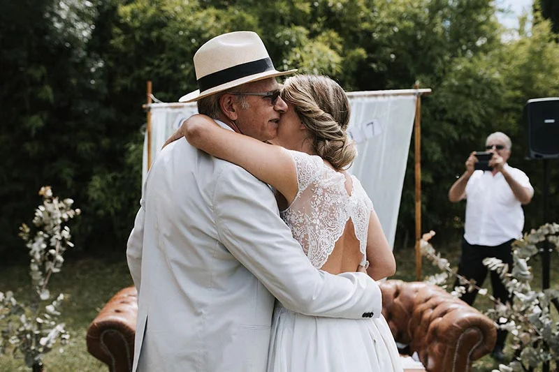 Une mariée en robe de dentelle embrasse tendrement un homme en costume blanc et chapeau lors d'une cérémonie en plein air.
