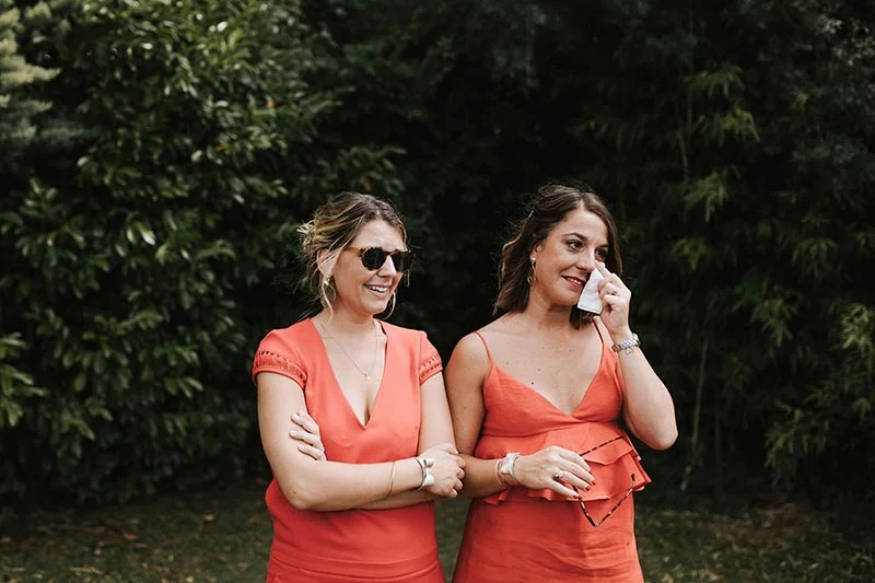 Deux femmes souriantes en robes corail partagent un moment d'émotion dans un jardin verdoyant.