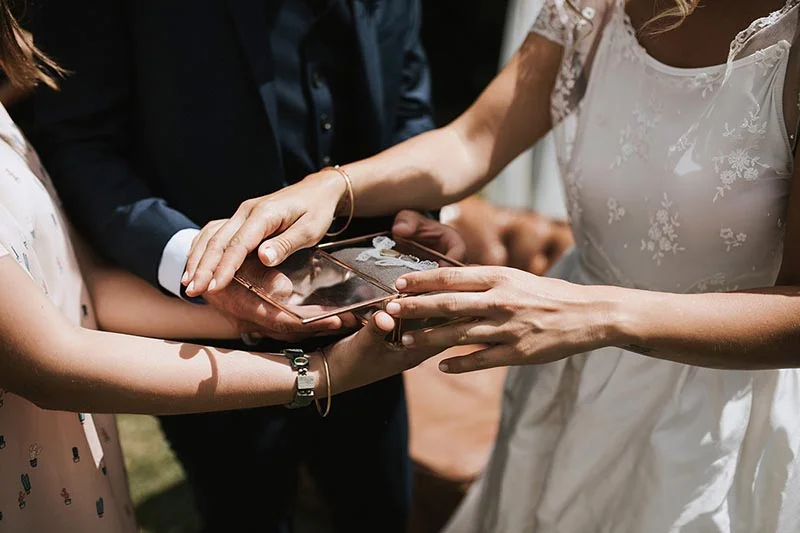 Gros plan sur les mains des mariés et d'une demoiselle d'honneur autour d'un écrin d'alliances lors d'un mariage.