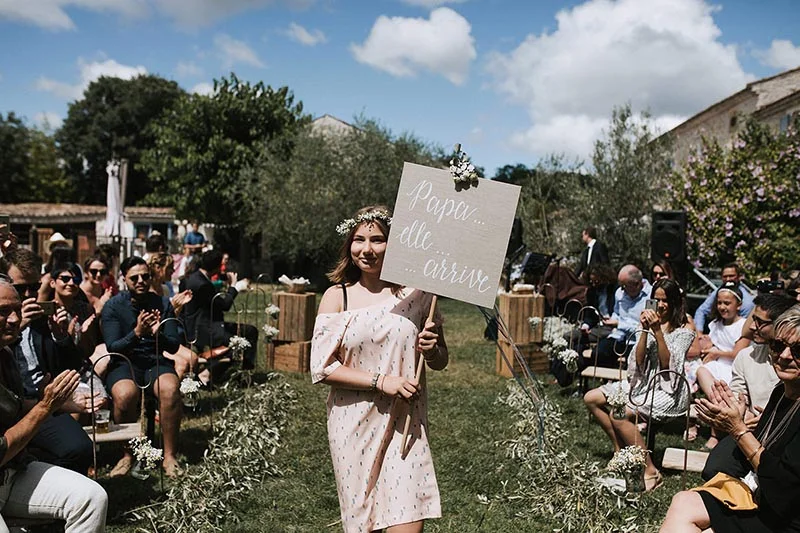 Une jeune fille en robe rose portant un panneau Papa arrive lors d'une cérémonie de mariage en plein air.