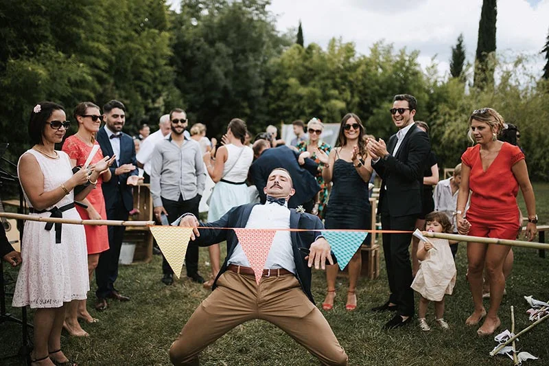 Invités souriants lors d'un mariage en extérieur, entourant un homme amusé sous une guirlande de fanions colorés.