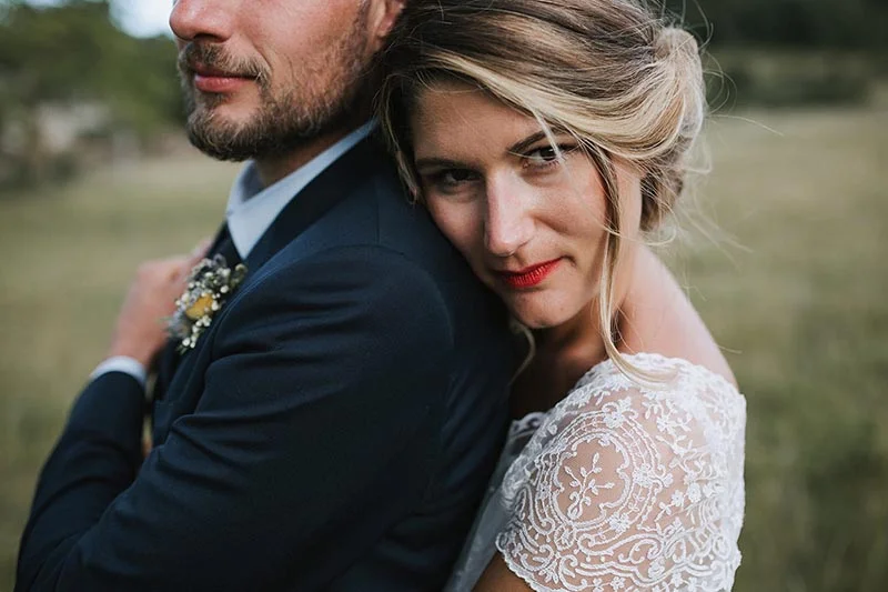 Jeune mariée en robe de dentelle blanche posant tendrement contre son époux en costume bleu marine.