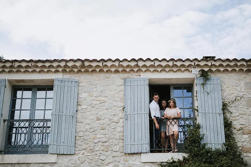 Trois personnes élégantes posent sur le balcon d'une maison en pierre aux volets bleu clair sous un ciel nuageux.