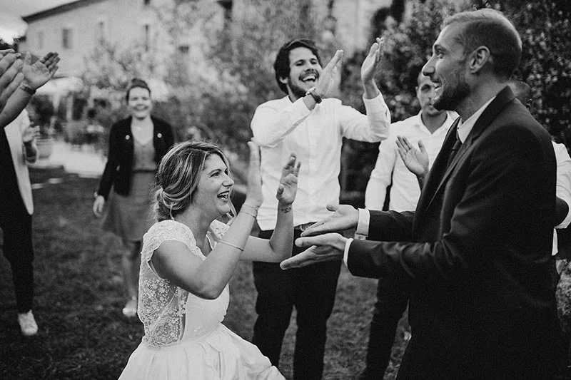 Couple de mariés joyeux entouré d'invités applaudissant en extérieur, photographie en noir et blanc.