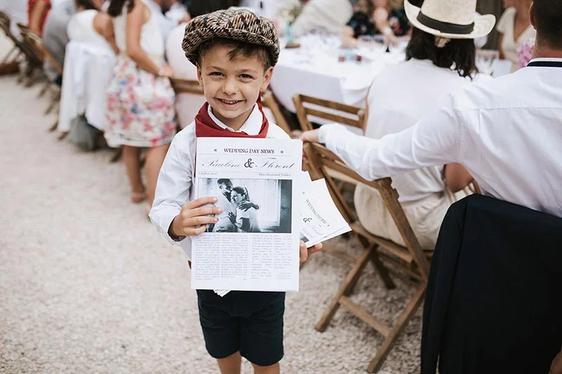 Un petit garçon souriant avec un béret tenant le journal du mariage de Pauline et Florent lors d'une réception en plein air.