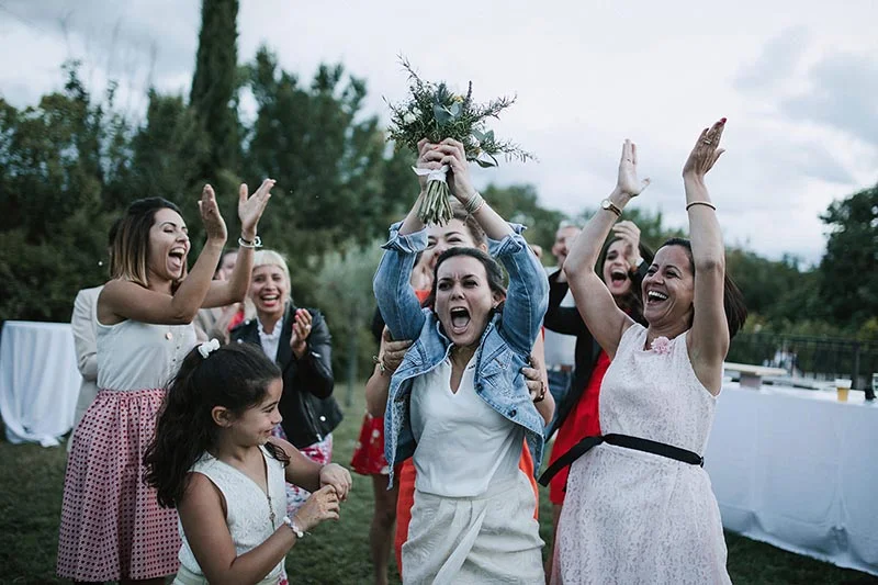 Une mariée et ses amies célèbrent avec joie en plein air, levant les bras et un bouquet dans une ambiance festive.
