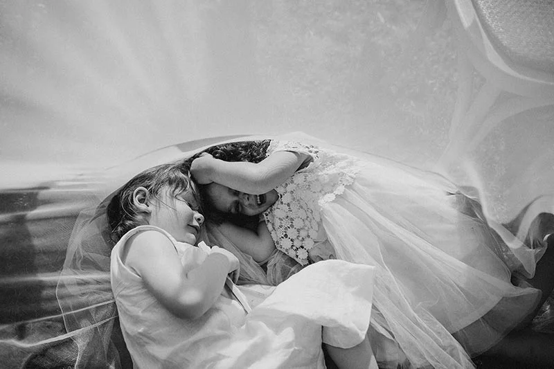 Deux petites filles endormies sous un voile de mariée blanc, photographie en noir et blanc à l'ambiance douce.