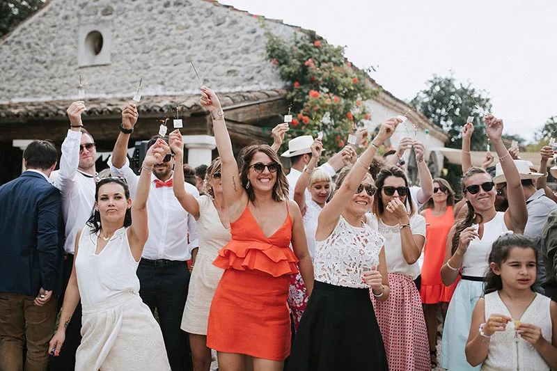 Invités souriants célébrant un mariage en extérieur en lançant des confettis devant une maison en pierre.