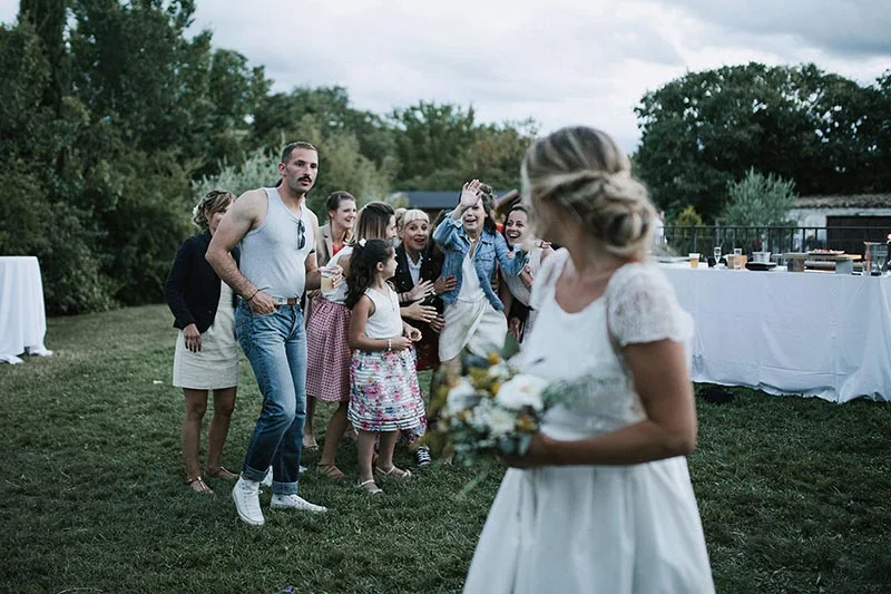 Une mariée de dos avec son bouquet face à des invités souriants lors d'une réception champêtre en extérieur.