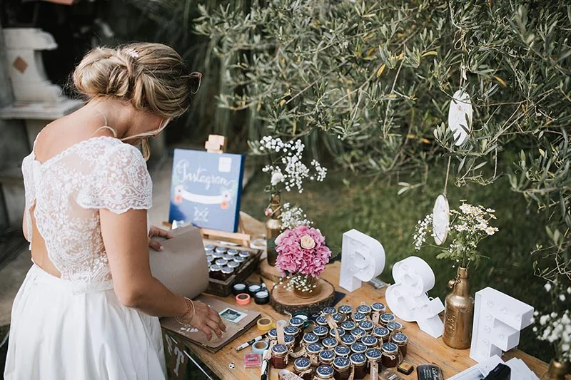 Une mariée en robe blanche prépare une table de cadeaux rustique avec des petits pots de confiture et des fleurs.