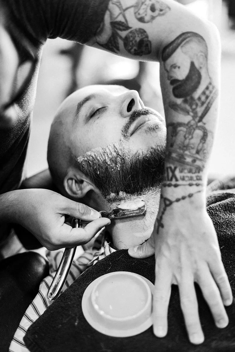 Photo noir et blanc d'un homme se faisant raser la barbe avec un coupe-chou par un barbier tatoué.