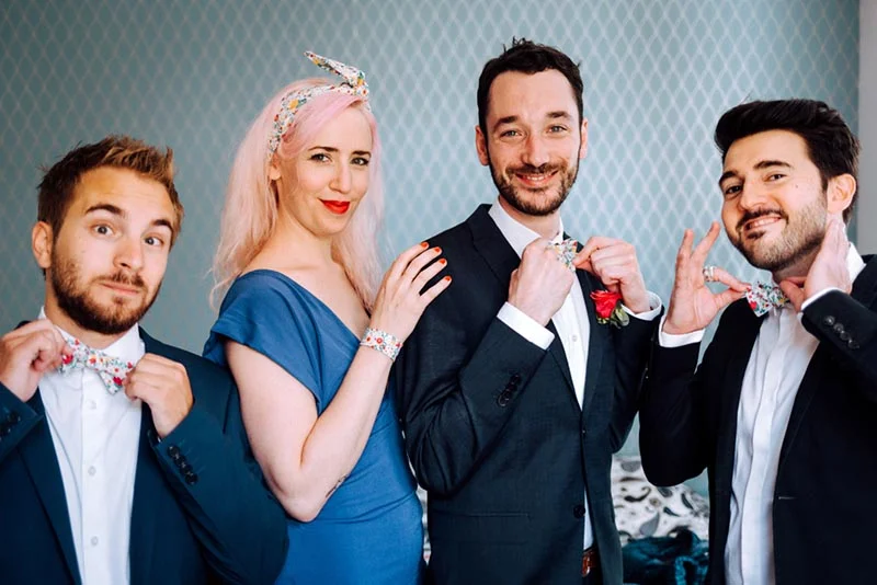 Quatre amis souriants, dont Mel et Ben, posant ensemble avec des accessoires floraux assortis sur fond bleu.