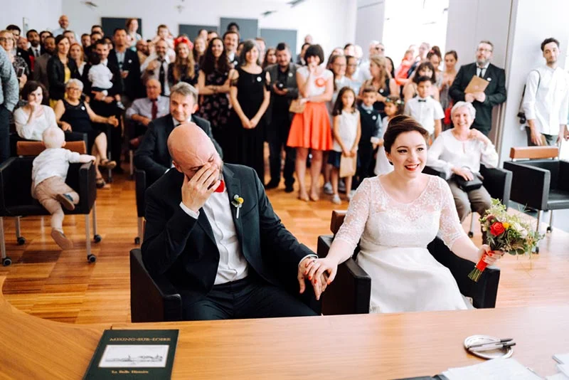 Un marié ému et sa femme rayonnante se tiennent la main lors de leur cérémonie civile entourés de leurs invités.