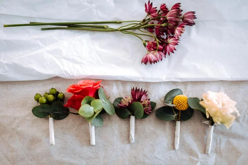 Boutonnières florales variées et fleurs rouges posées sur un papier blanc et kraft pour le mariage de Mel & Ben.