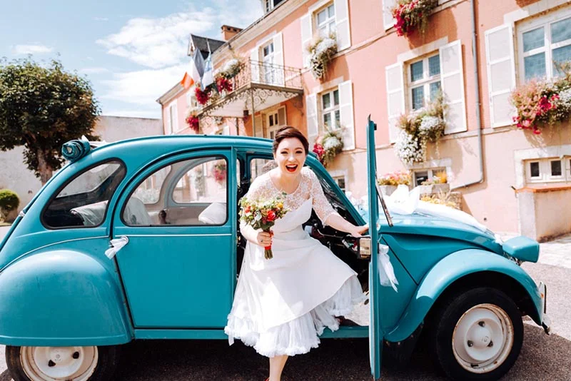 Une mariée souriante avec un bouquet sort d'une Citroën 2CV bleue devant des maisons roses au style provençal.