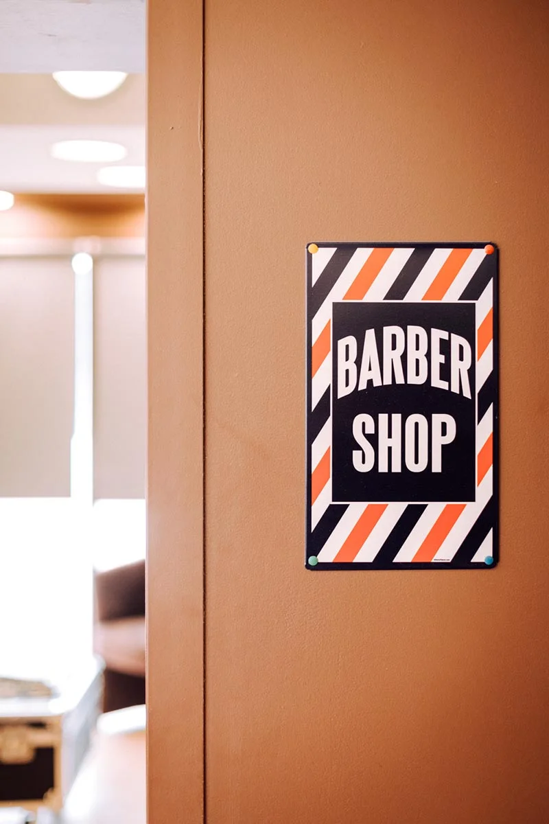 Plaque signalétique vintage d'un barber shop aux rayures orange, noires et blanches sur un mur beige.