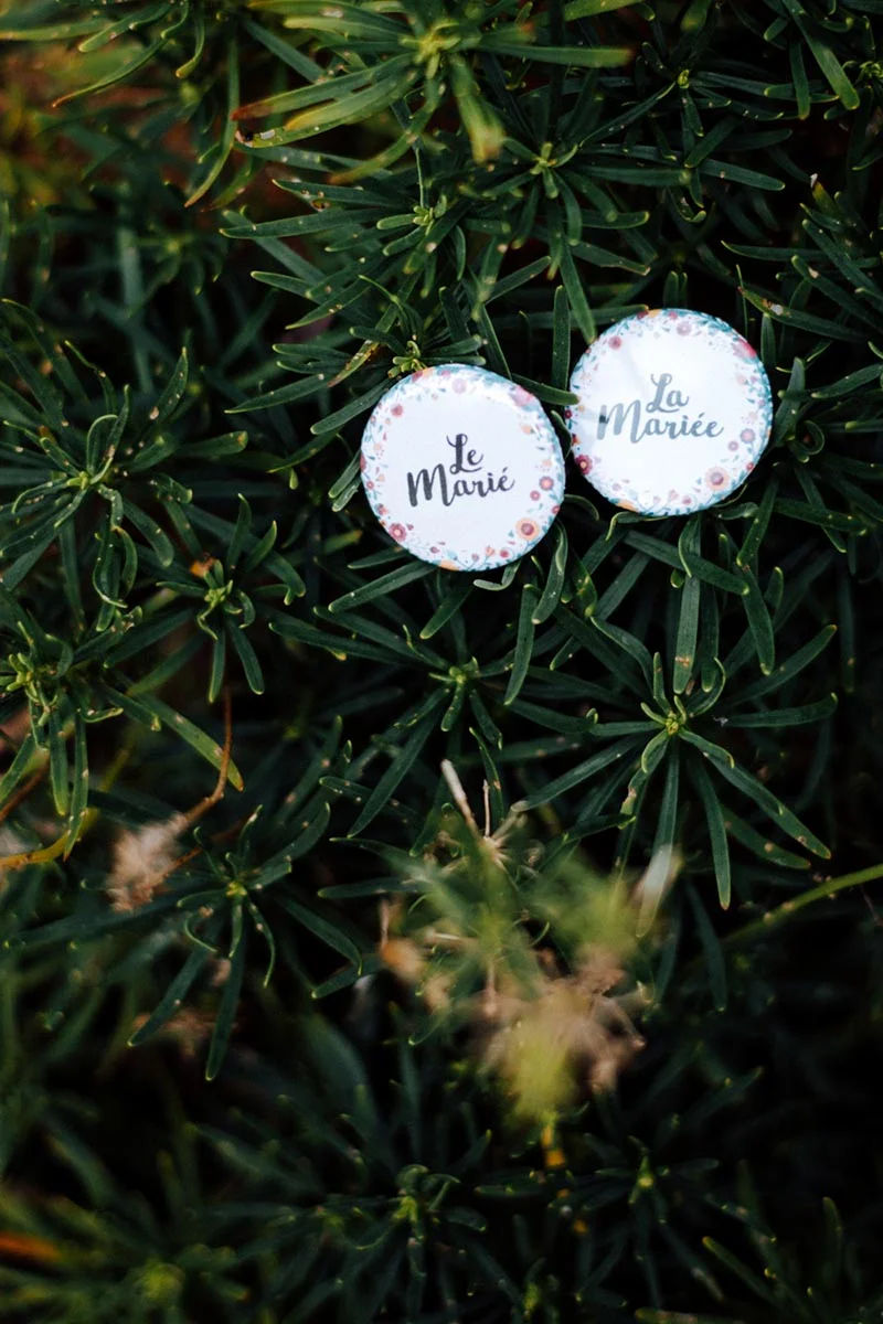 Deux badges ronds blancs avec inscriptions Le Marié et La Mariée, posés sur un feuillage vert dense.