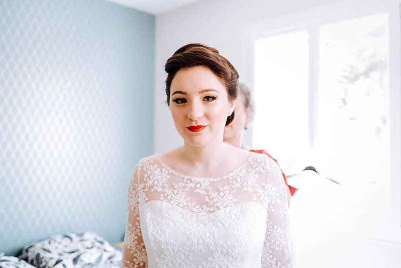 Une mariée au maquillage sophistiqué et robe en dentelle blanche pose dans une pièce lumineuse.