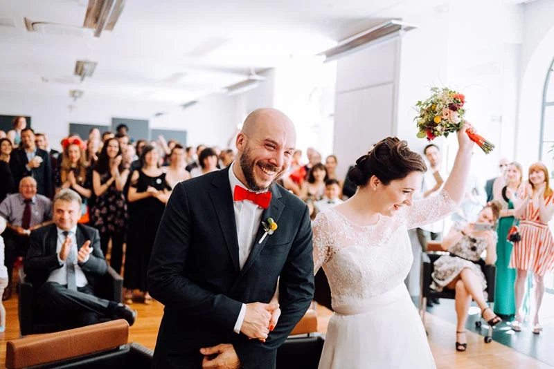 Jeunes mariés souriants, lui en costume noir et nœud papillon rouge, elle en robe blanche, devant leurs invités.