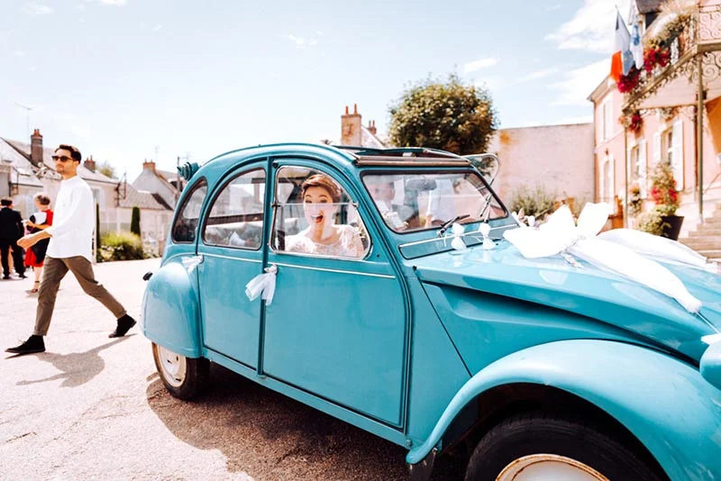 Une mariée souriante dans une Citroën 2CV bleue décorée de rubans blancs dans un village français.