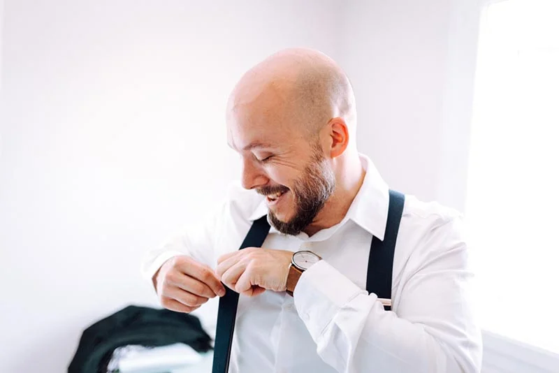 Un marié chauve et barbu en chemise blanche et bretelles noires s'apprête avec un sourire serein.