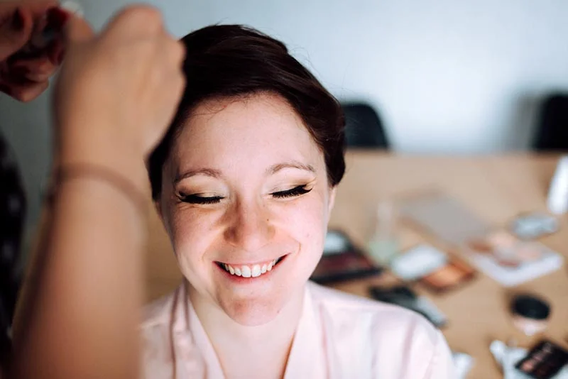 Une mariée souriante se fait coiffer et maquiller dans des tons doux et lumineux.