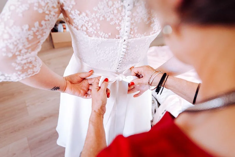 Une main aide une mariée à nouer le ruban blanc de sa robe en dentelle.