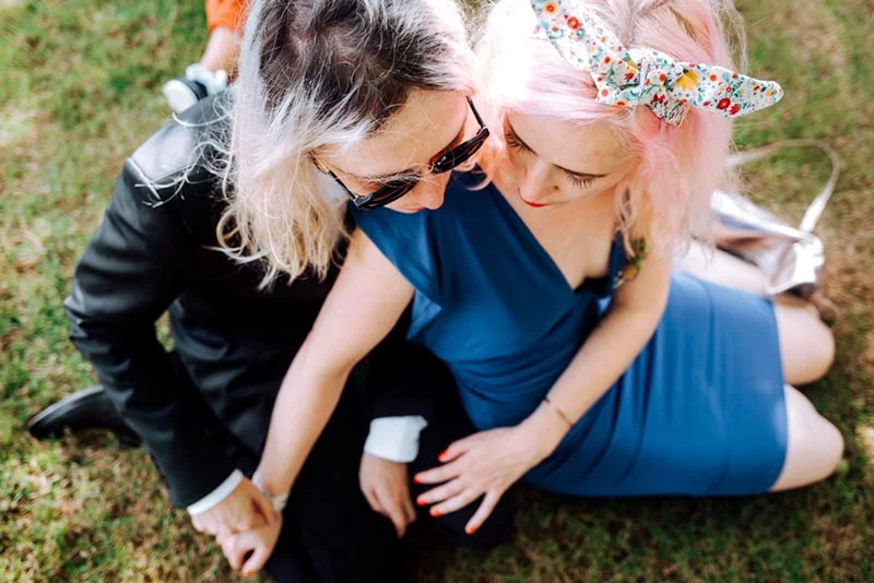 Couple complice, l'une en robe bleue et cheveux roses, l'autre en veste noire, s'enlaçant tendrement sur l'herbe.