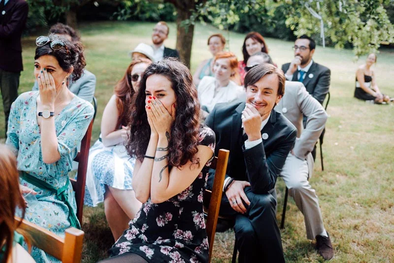 Invités émus et souriants assistant à la cérémonie de Mel et Ben en plein air dans un jardin verdoyant.