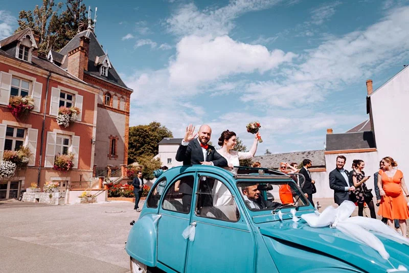 Jeunes mariés souriants dans une Citroën 2CV bleue devant un bâtiment ancien, entourés d'invités sous un ciel bleu.
