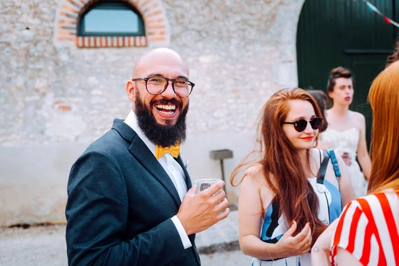 Un homme souriant en costume et nœud papillon jaune discute avec des invités lors d'un mariage en extérieur.