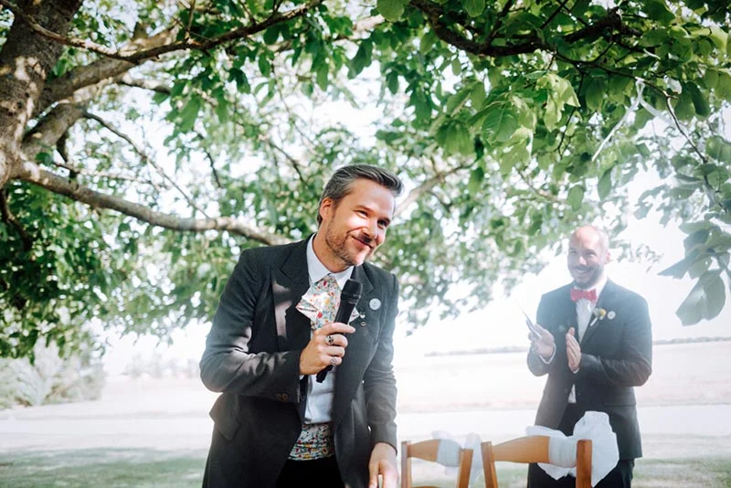 Un homme en costume gris et nœud papillon coloré prononce un discours souriant lors d'une cérémonie en plein air.